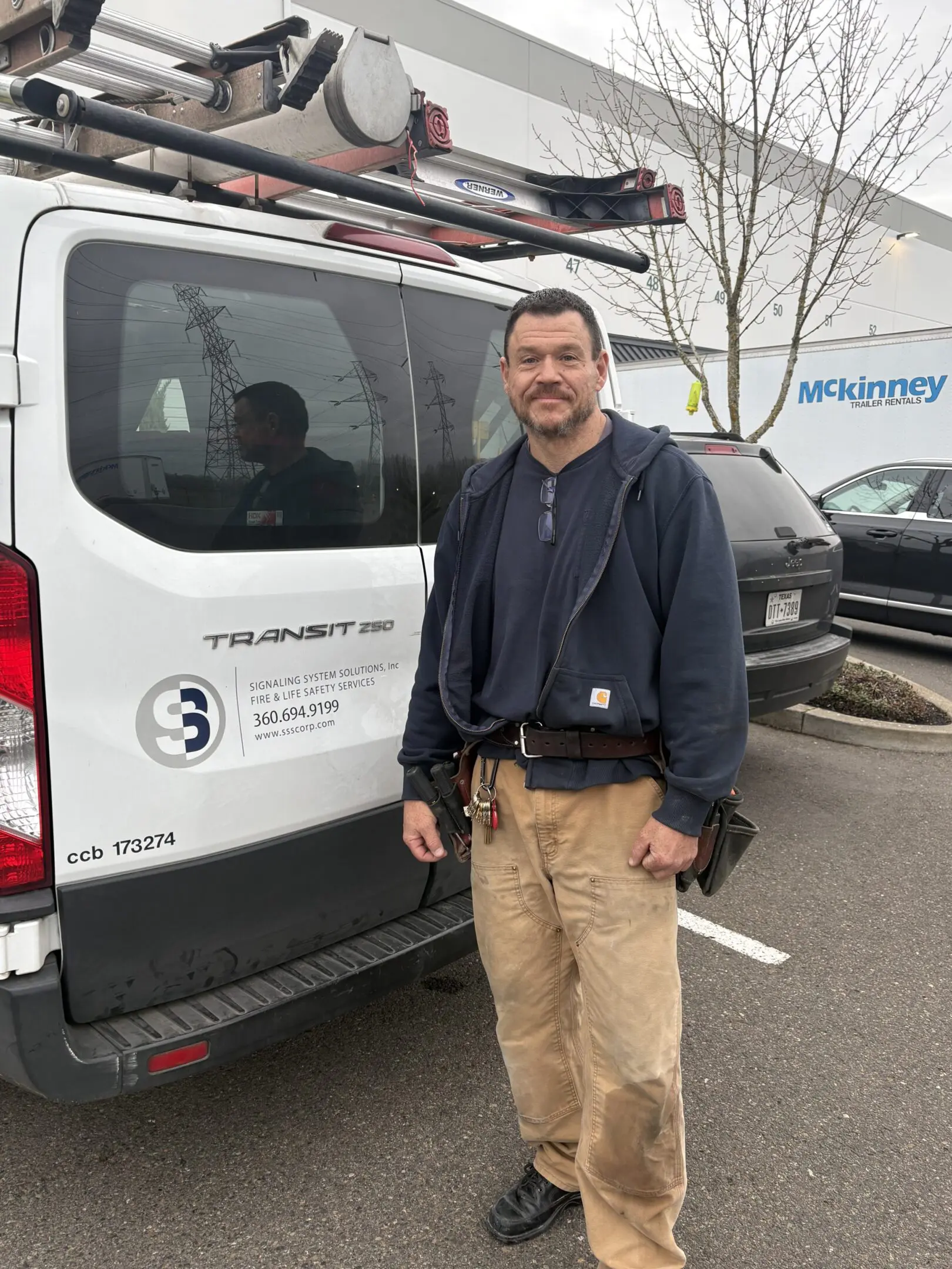 Man standing next to a white work van with tools in his pocket.