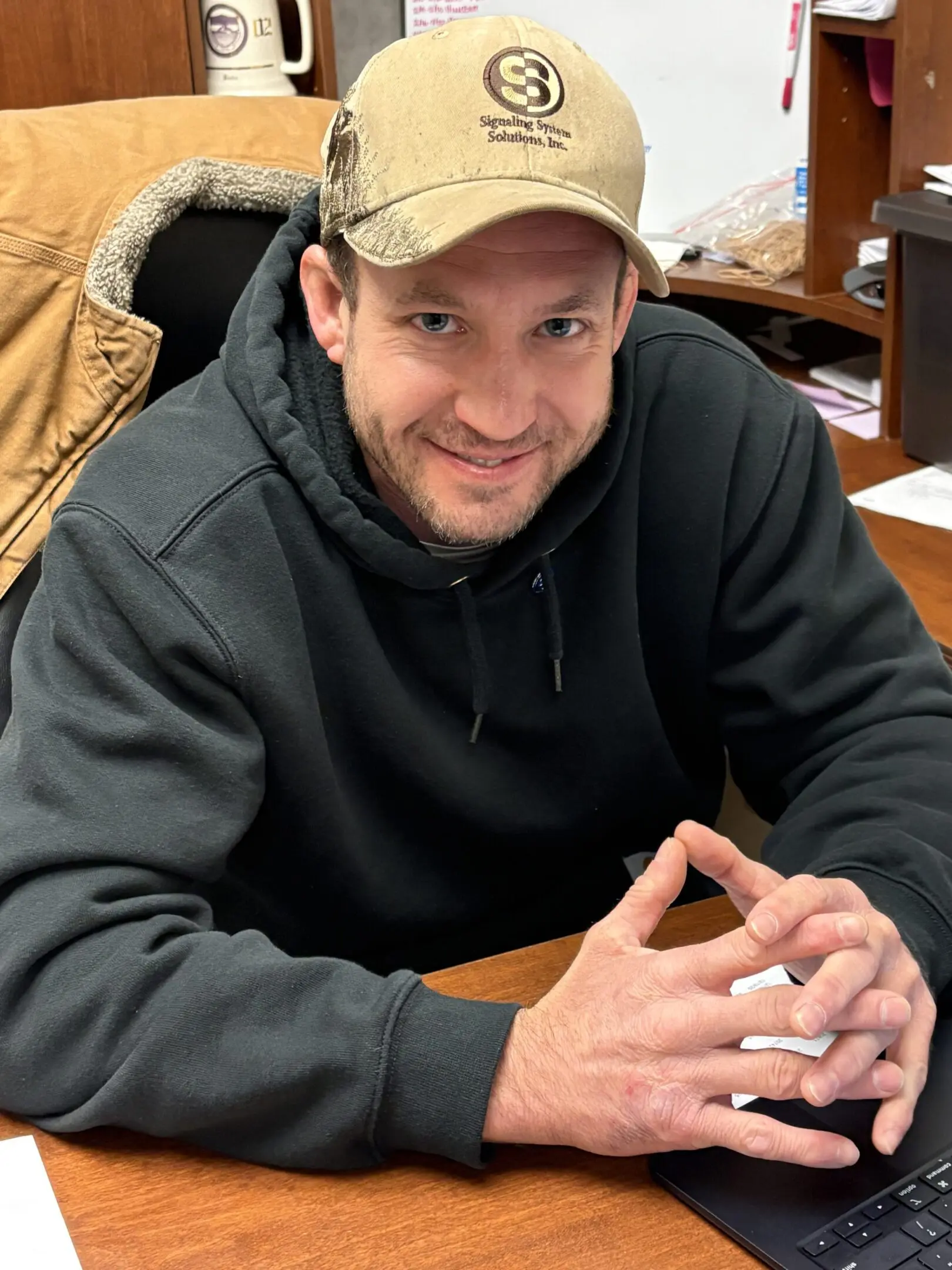Smiling man in a black hoodie and cap sitting at a desk indoors.