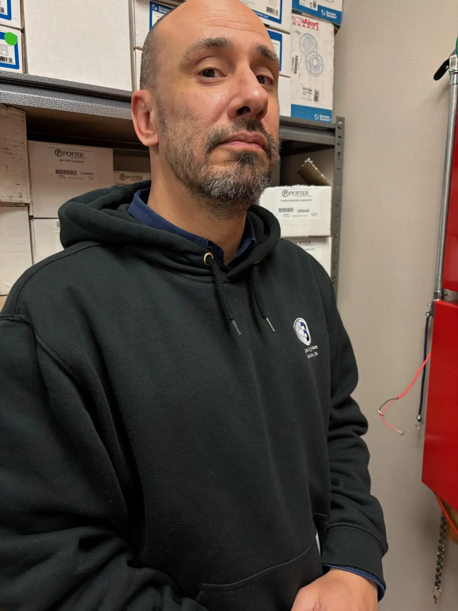 Man in black hoodie standing indoors near shelves and a red structure.