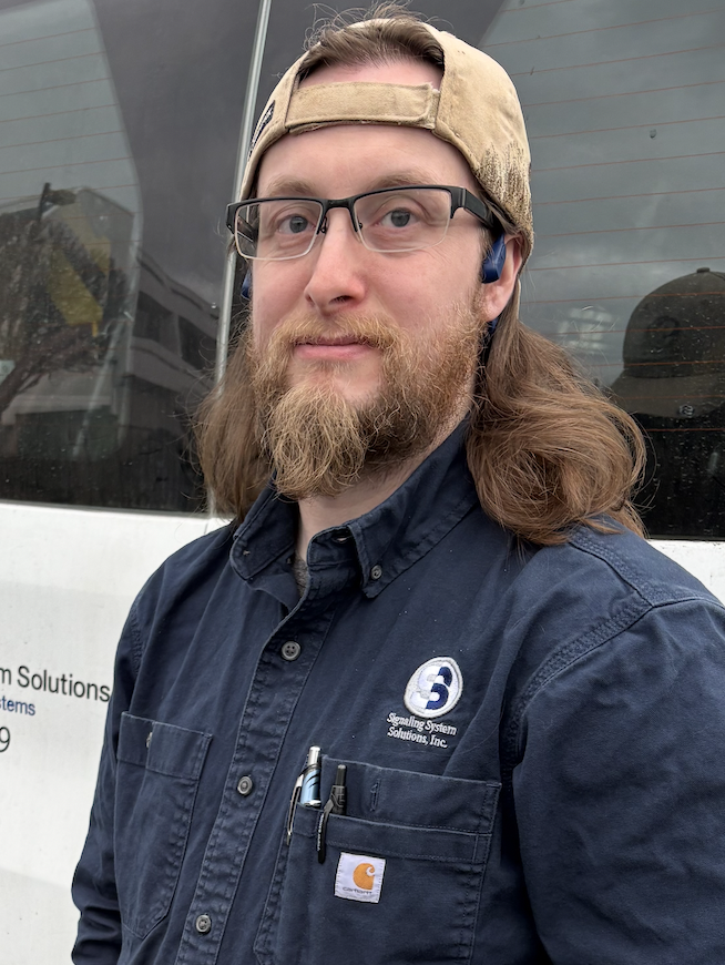 Man with glasses and beard wearing a cap and navy work shirt.